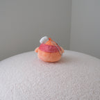 A small peach-colored plush toy with a red-and-white gingham scarf and a white hat sits on a round, textured white pouf against a plain gray wall.