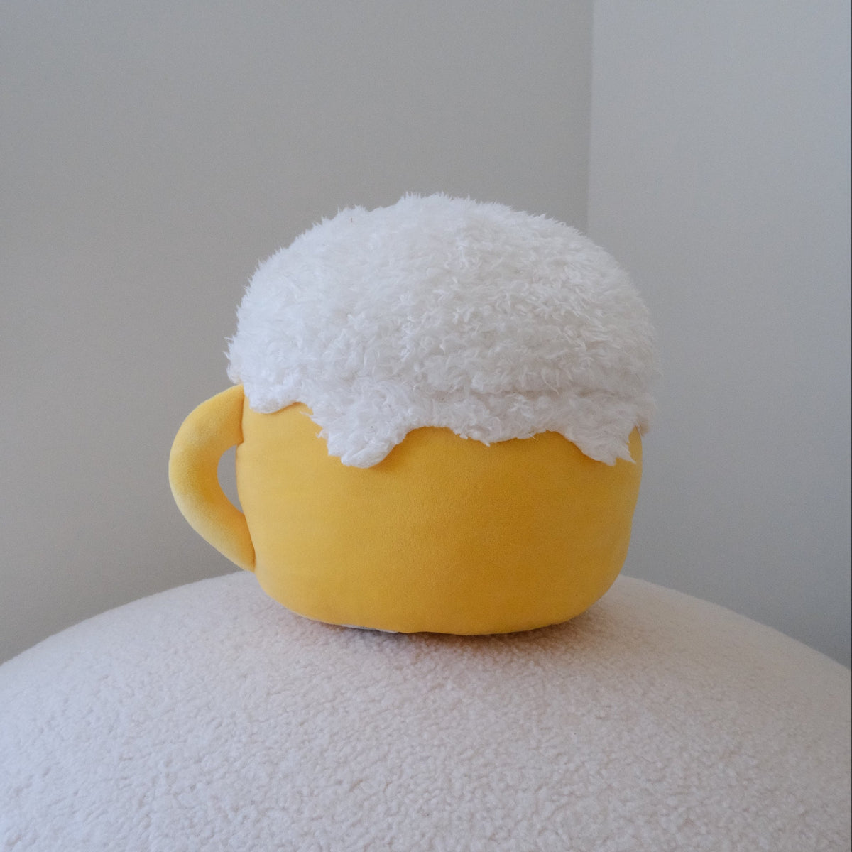 A yellow mug-shaped plush with a white fluffy faux-fur top, resting on a textured white surface against a plain light gray background.