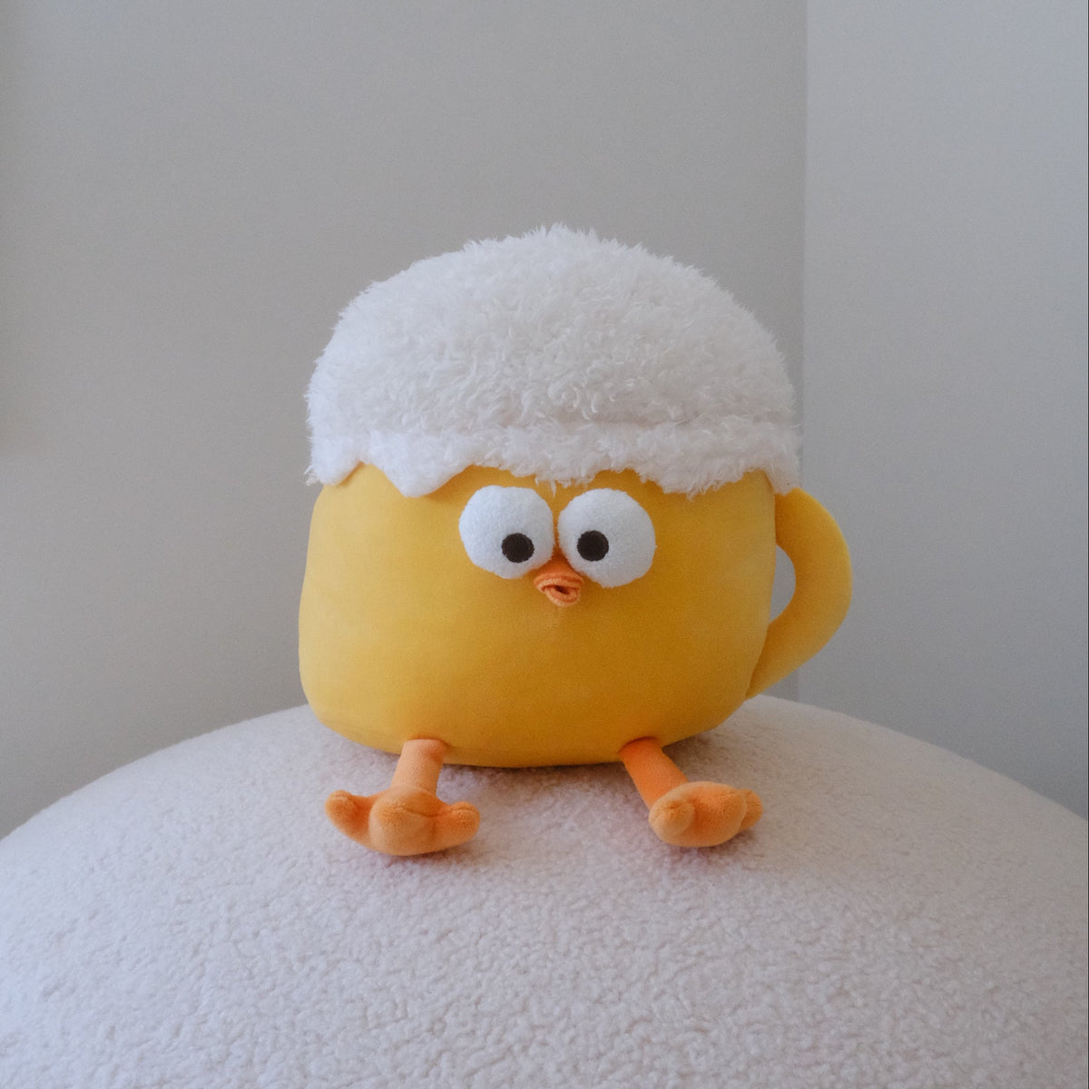A yellow plush toy shaped like a chicken shaped like white fuzzy mug, with orange feet and hands, posed on a white textured surface against a plain light gray wall.