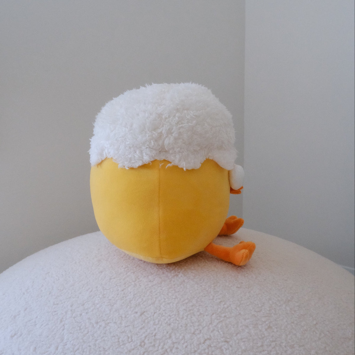 A yellow plush toy with a white fluffy top and orange feet, sitting on a white textured surface against a plain light gray wall.