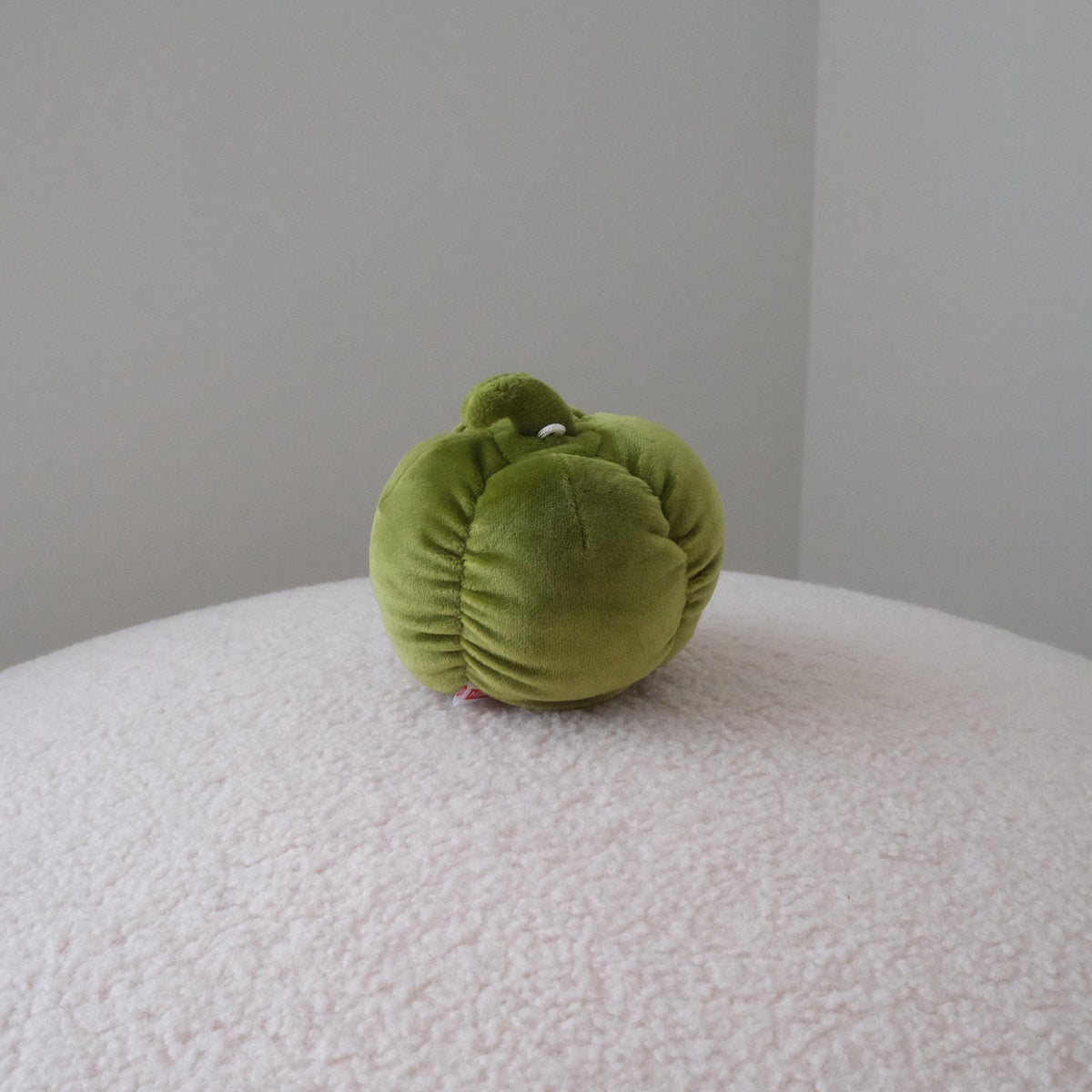 A small olive green plush bell pepper toy with a textured velvet-like fabric, sitting on a white textured surface against a neutral gray background.