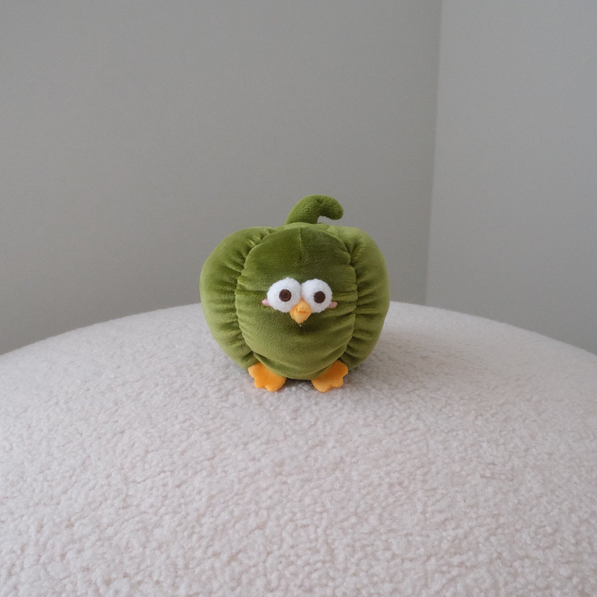 A small green bell pepper plush toy with a curly stem, large white eyes, and orange feet, sitting on a textured cream-colored surface against a plain gray wall.