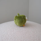A small green bell pepper plush toy with orange feet and a white side detail sits on a round, textured white ottoman against a plain gray wall.