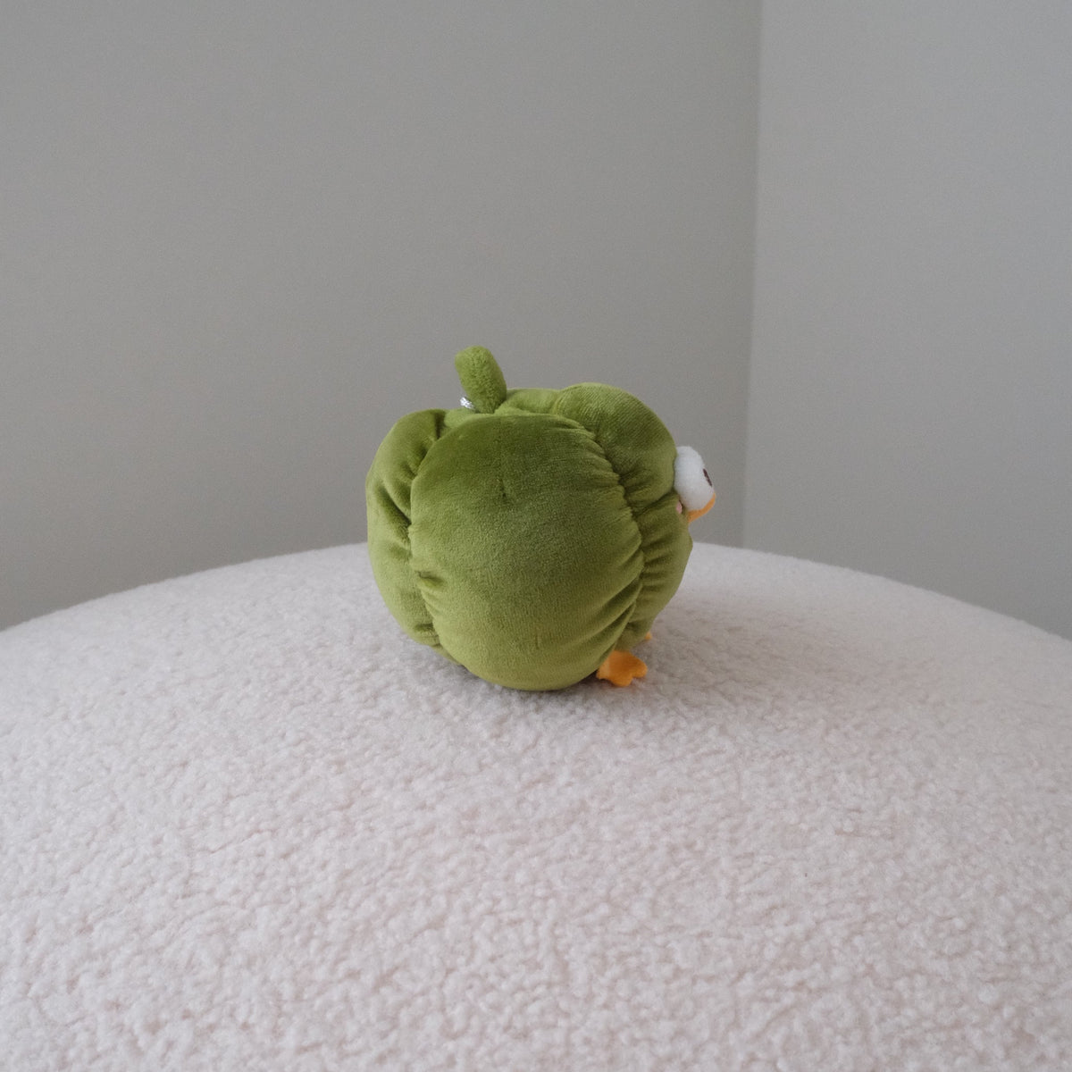 A small green bell pepper plush toy with orange feet and a white side detail sits on a round, textured white ottoman against a plain gray wall.