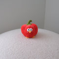 A small red plush toy shaped like a bell pepper with a green stem and embroidered face, standing on a white textured surface against a plain gray wall.