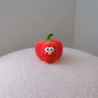 A small red plush toy shaped like a bell pepper with a green stem and embroidered face, standing on a white textured surface against a plain gray wall.