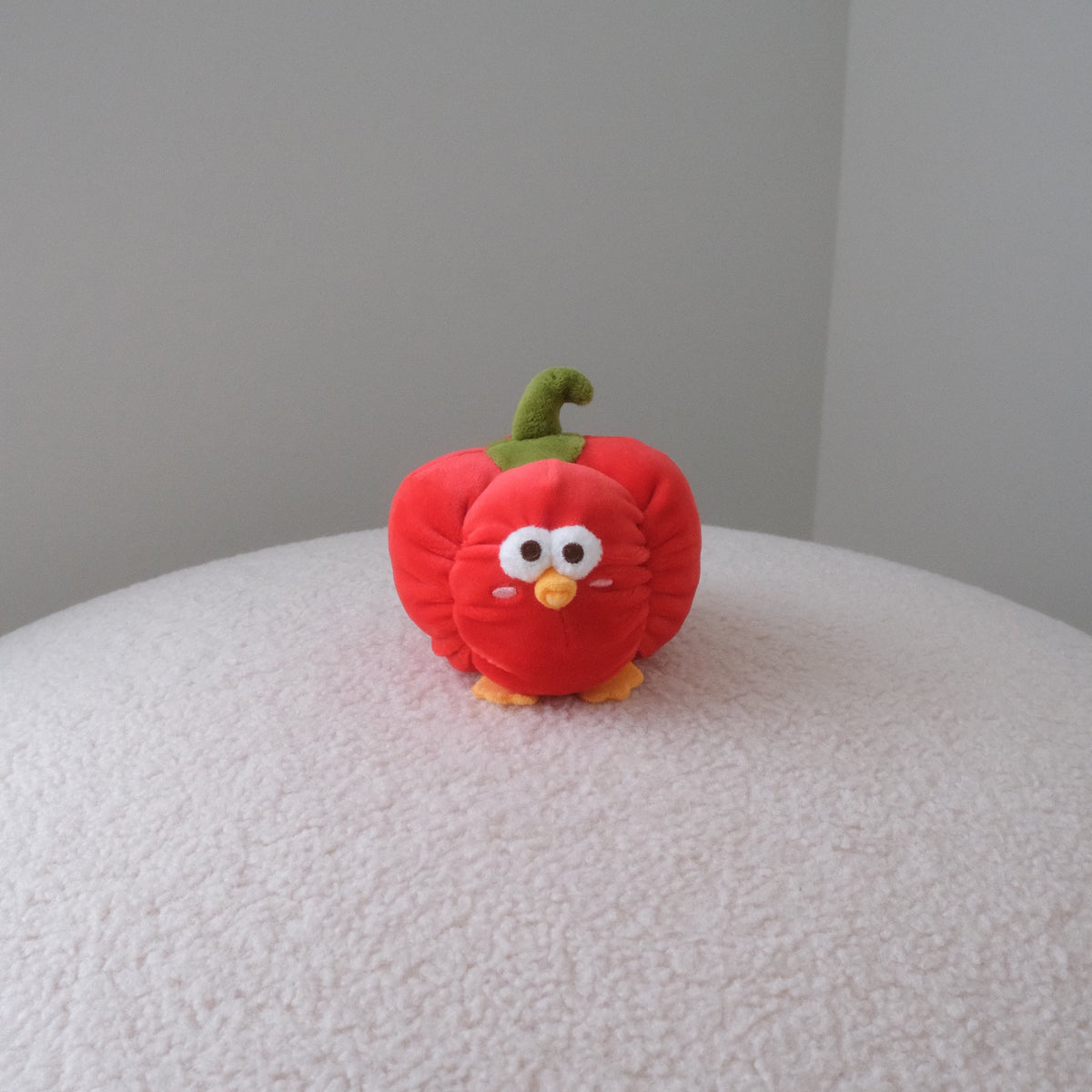 A small red plush toy shaped like a bell pepper with a green stem and embroidered face, standing on a white textured surface against a plain gray wall.