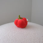 A small red plush bell pepper toy with a green stem and a tiny white accent, sitting on a textured white surface against a plain gray wall.