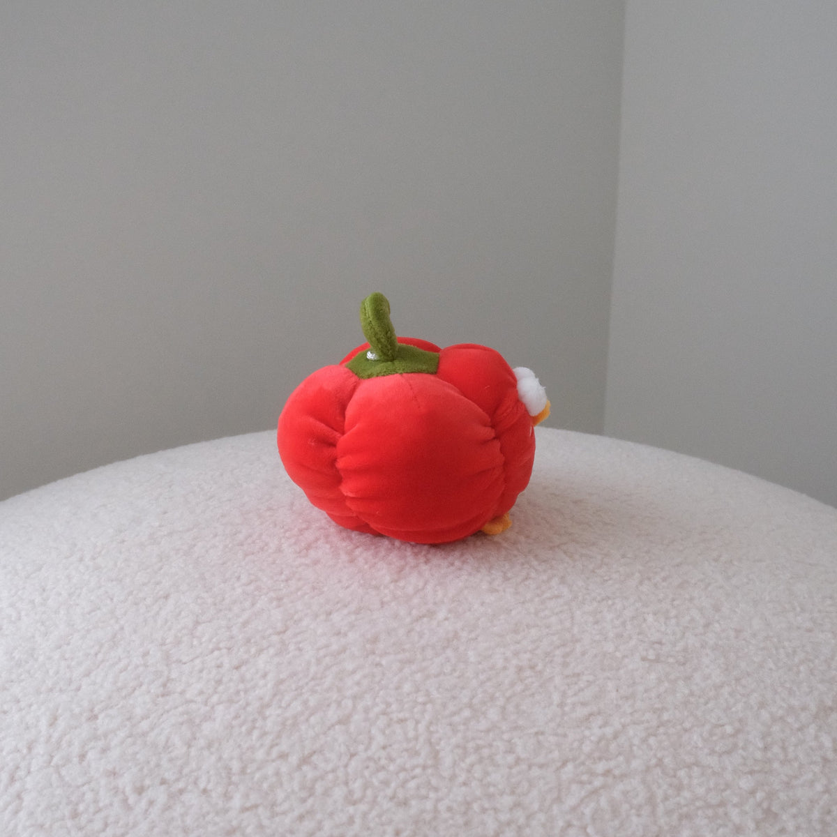 A small red plush bell pepper toy with a green stem and a tiny white accent, sitting on a textured white surface against a plain gray wall.