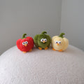 Three small plush vegetable characters resembling a red pepper, green pepper, and yellow pepper, standing on a textured white surface against a plain light gray background.