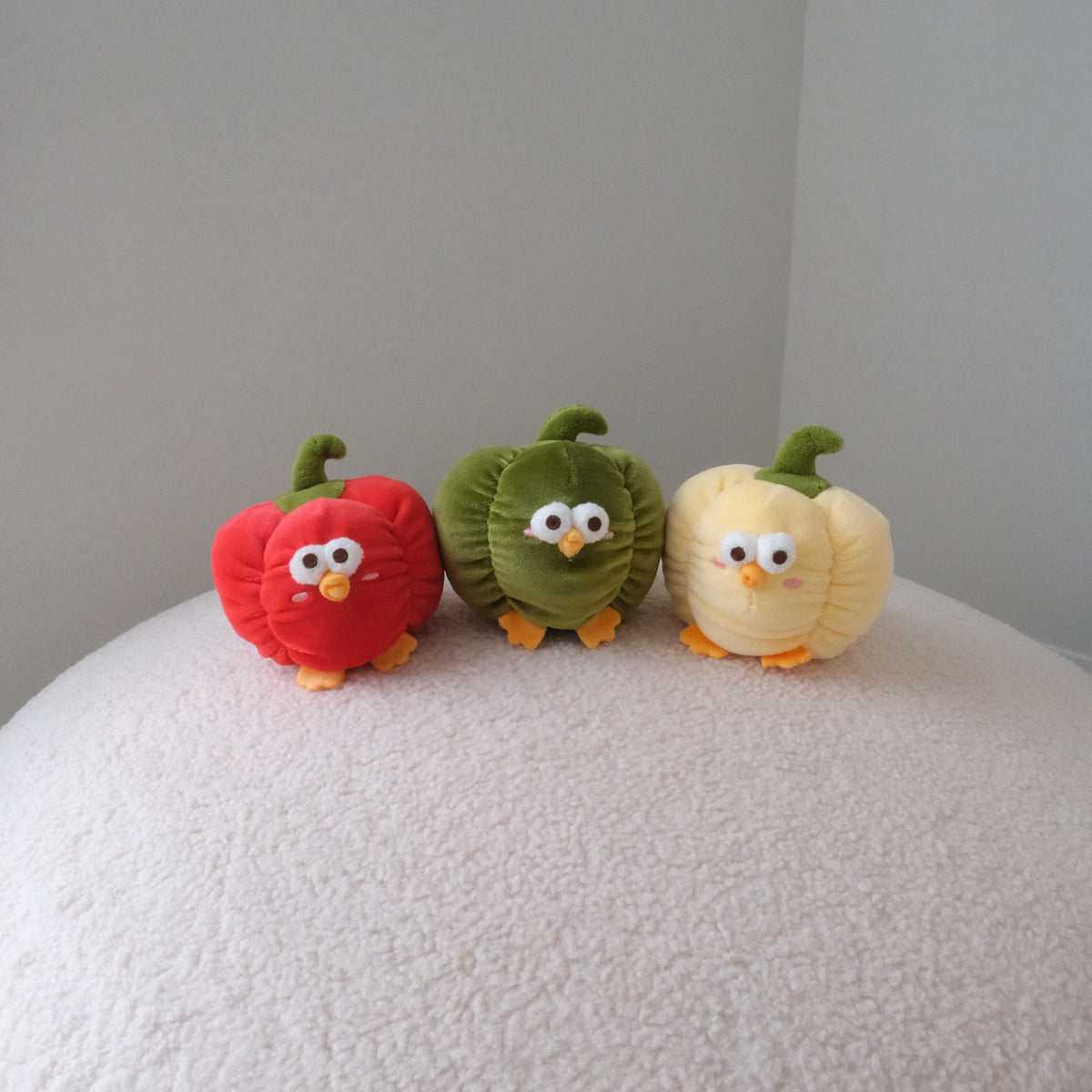 Three small plush vegetable characters resembling a red pepper, green pepper, and yellow pepper, standing on a textured white surface against a plain light gray background.