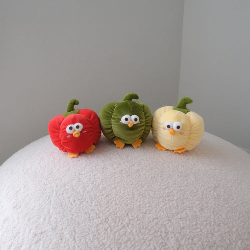 Three small plush vegetable characters resembling a red pepper, green pepper, and yellow pepper, standing on a textured white surface against a plain light gray background.