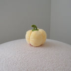 A small pale yellow plush bell pepper toy with a green stem sits on a textured white surface against a plain gray background.