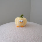 A small yellow plush bell pepper toy with a green stem and orange feet, standing on a textured white surface against a plain gray wall.