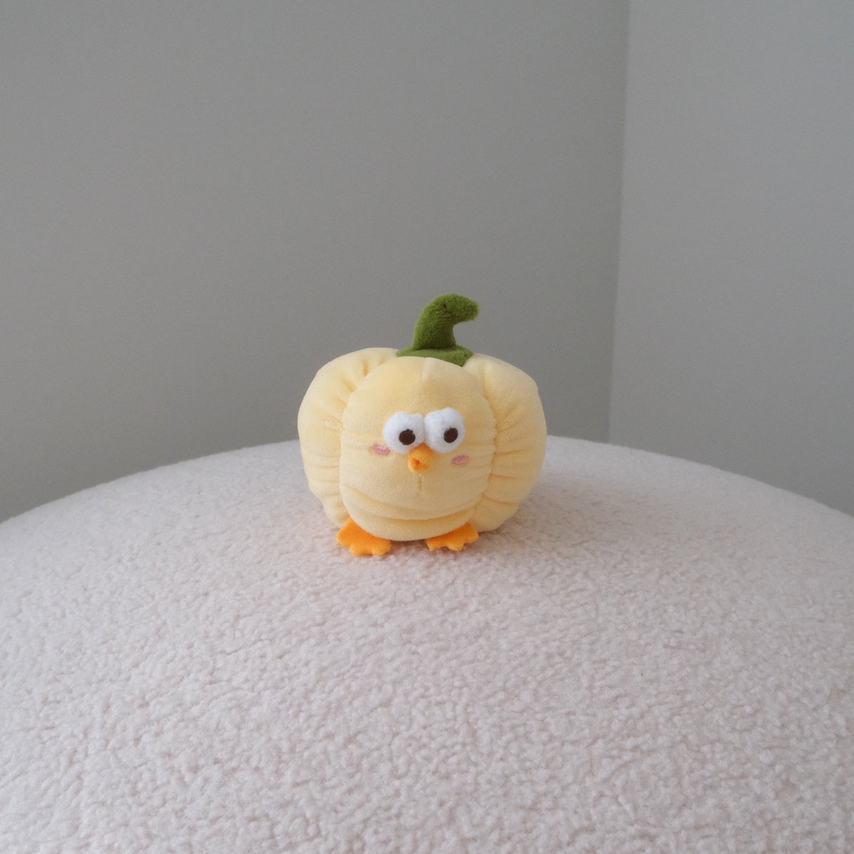 A small yellow plush bell pepper toy with a green stem and orange feet, standing on a textured white surface against a plain gray wall.