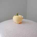 A small yellow plush bell pepper toy with a green stem and orange feet sits on a textured white surface against a plain gray background.
