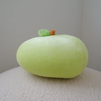 A light green apple plush toywith a small brown stem and green leaf, resting on a textured cream surface against a neutral gray background.