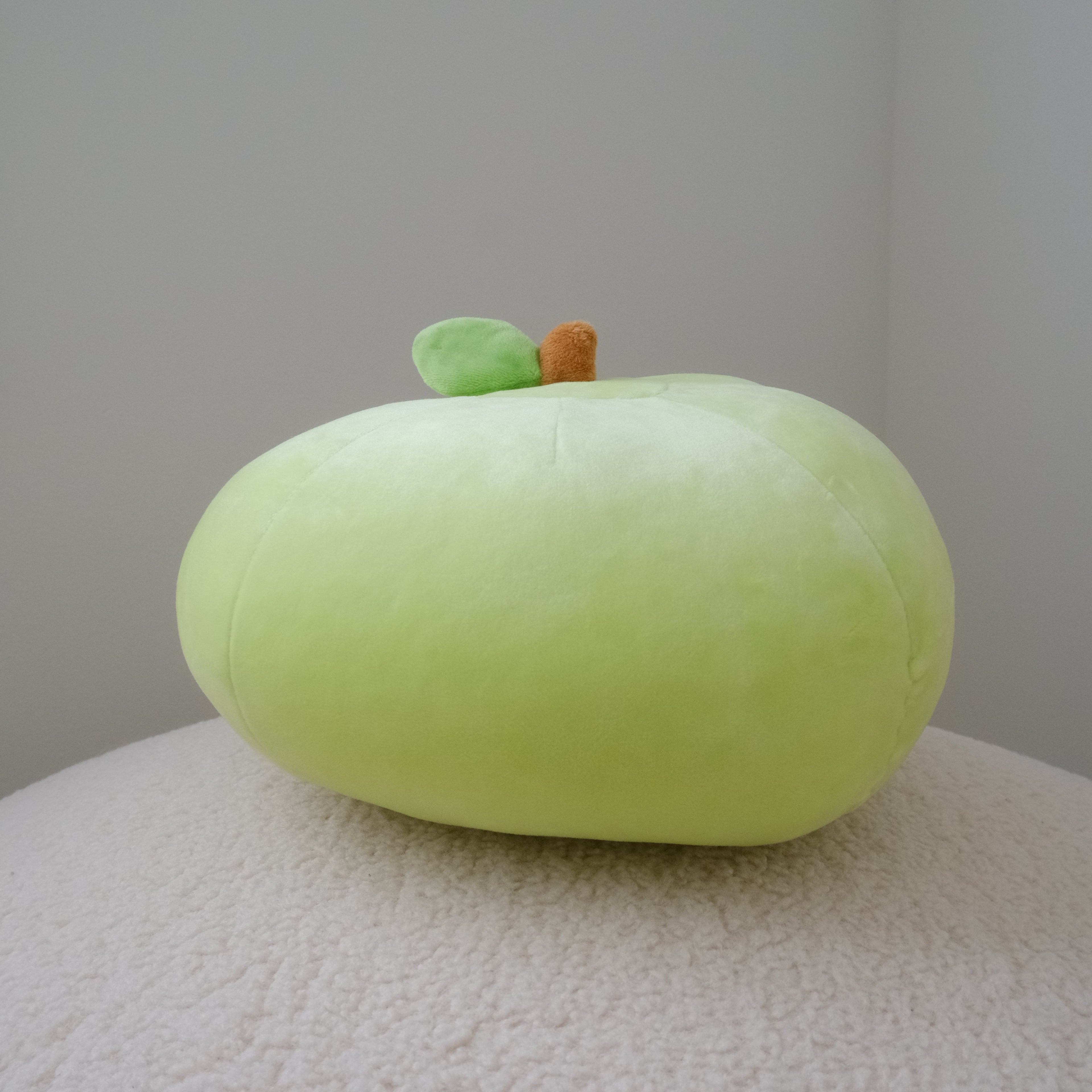 A light green apple plush toywith a small brown stem and green leaf, resting on a textured cream surface against a neutral gray background.