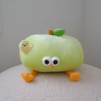 A cute, round green plush toy shaped like an apple, sitting on a textured white surface against a plain light gray wall. The plush has large cartoon eyes, orange legs, a small green leaf on top, and a small plush worm attached to its side.