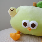 A close-up of a cute, soft plush toy shaped like a smiling light green apple, with large white eyes, a small orange beak, yellow feet, and a green leaf on top, resting on a white textured surface against a neutral gray background. A cute worm plush with a smiley face is coming out of the side of the apple.