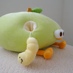 A plush green apple toy with a yellow worm dangling from its back, resting on a textured white surface against a neutral gray background.
