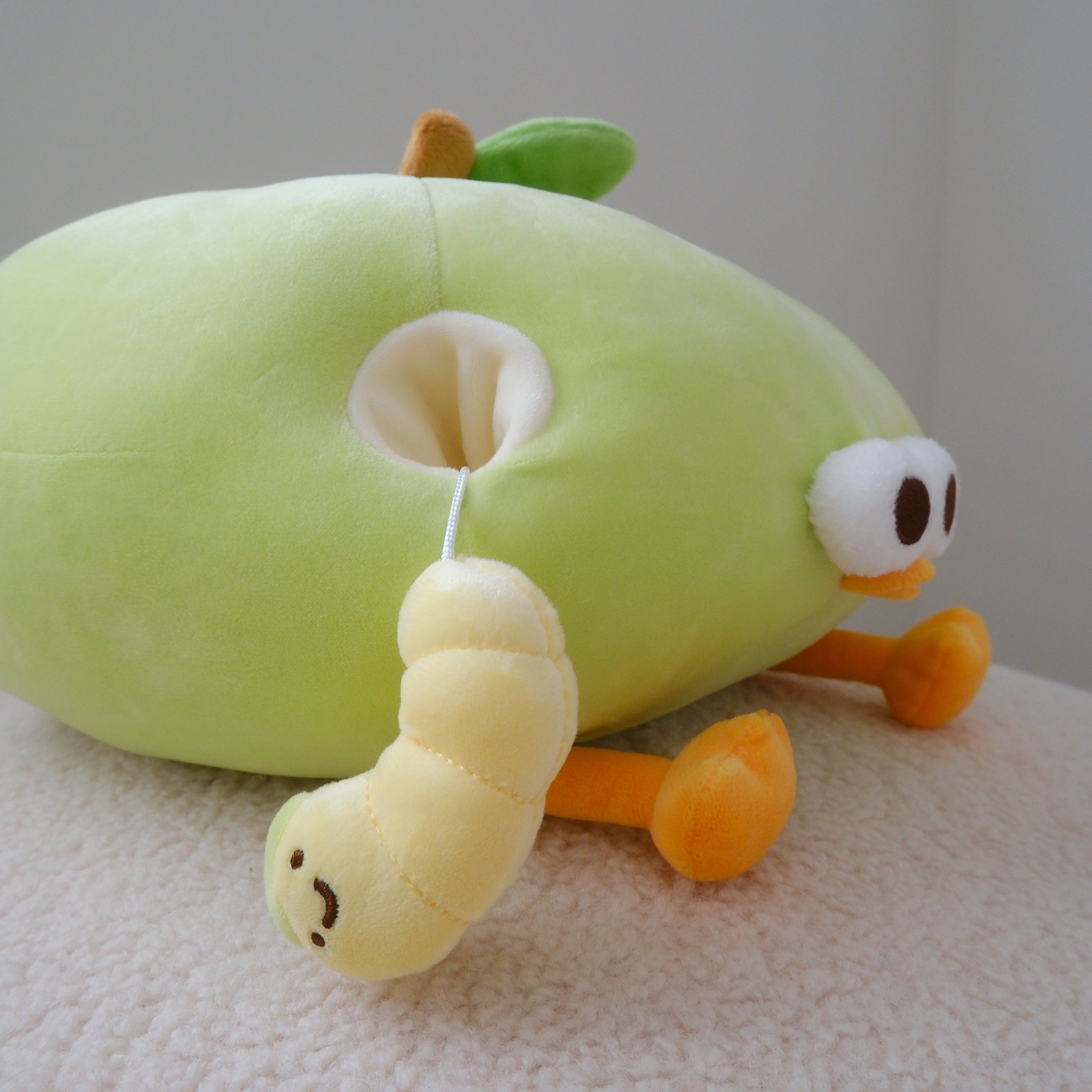 A plush green apple toy with a yellow worm dangling from its back, resting on a textured white surface against a neutral gray background.