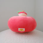 A red plush apple-shaped cushion with a small decorative topper on top, resting on a textured white surface against a plain light gray background.