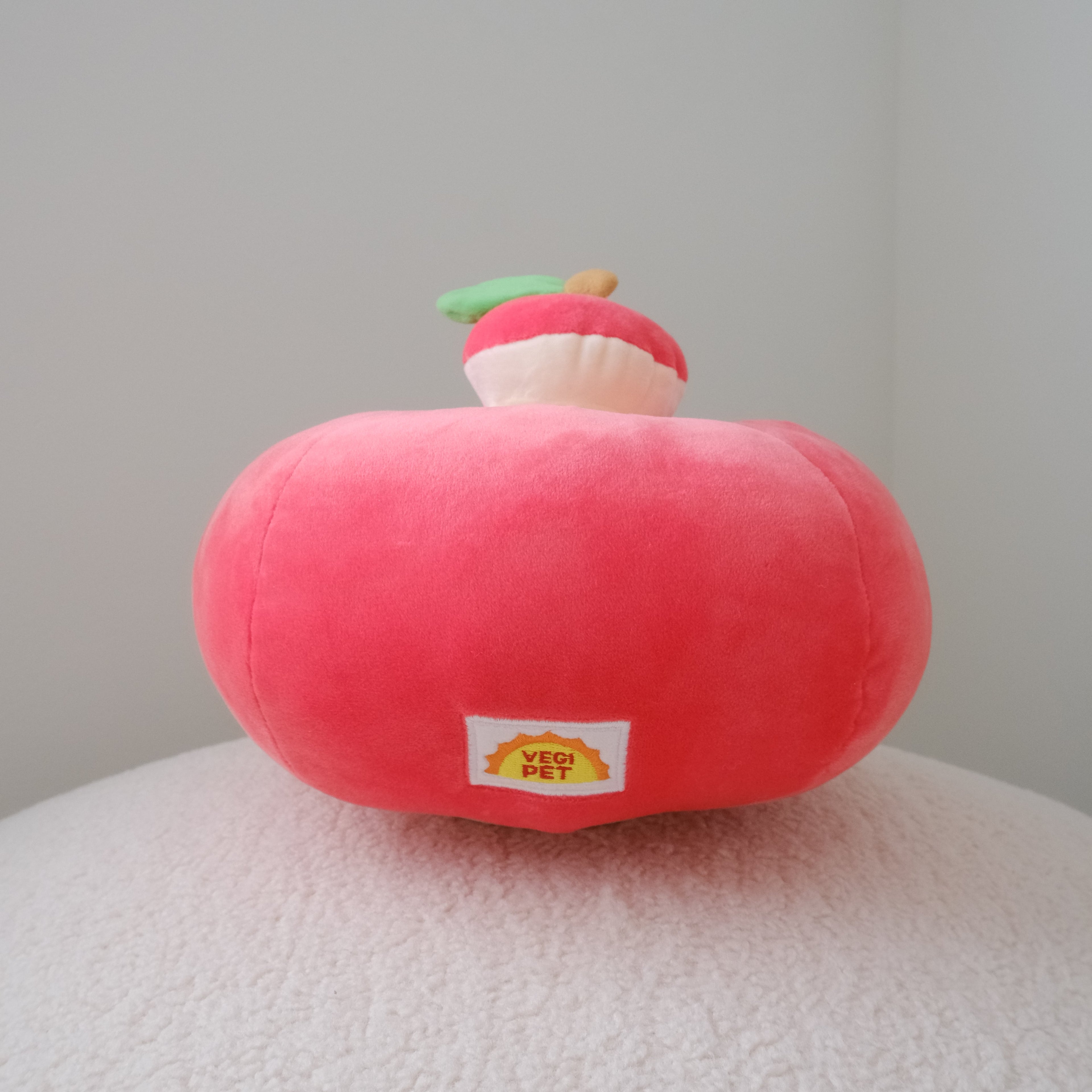 A red plush apple-shaped cushion with a small decorative topper on top, resting on a textured white surface against a plain light gray background.