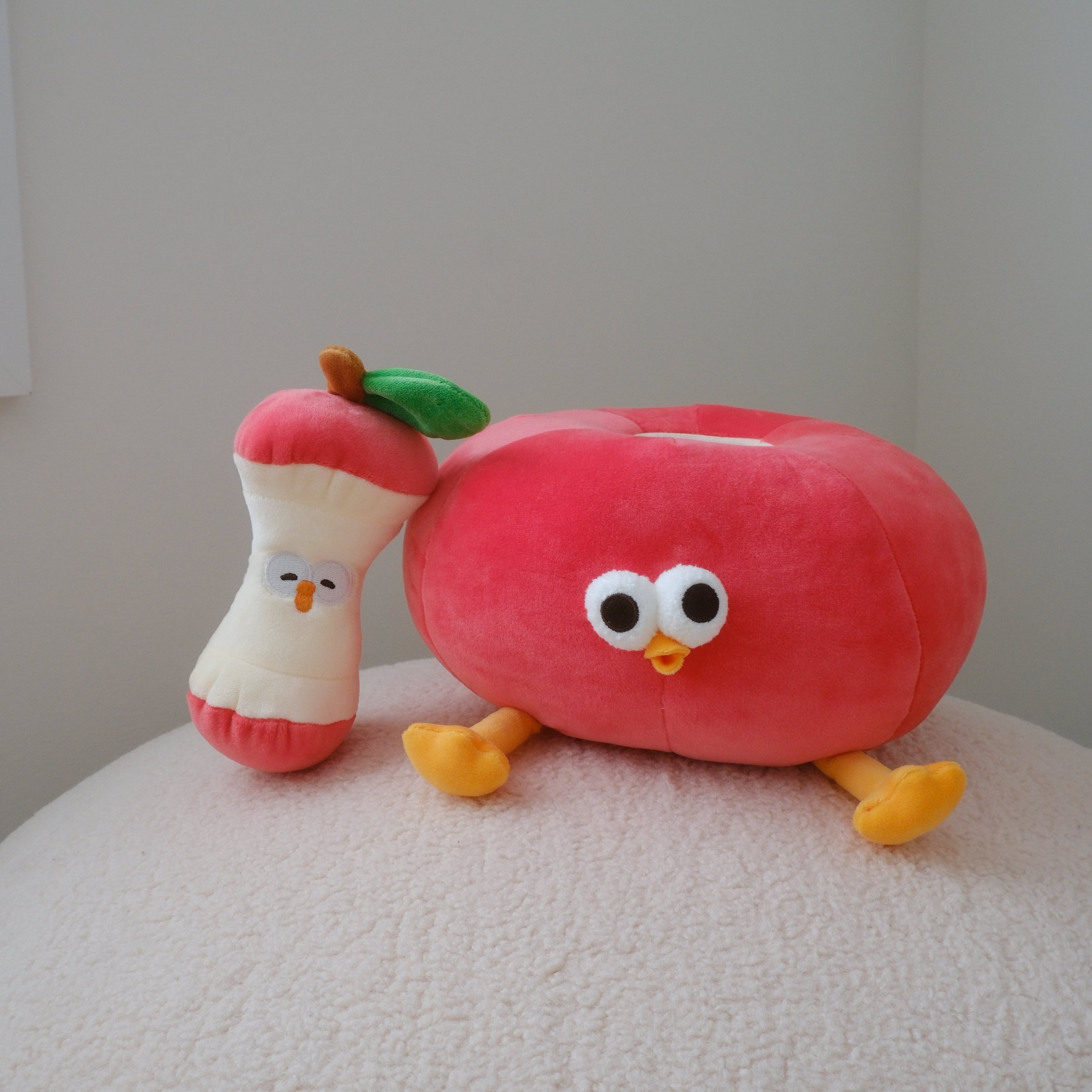 A plush red apple toy with a cartoon face and yellow legs sits on a textured white surface against a plain light gray wall. An apple core plush sits next to the apple.