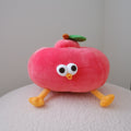 A red plush toy shaped like an apple with large white eyes, a small orange beak, yellow legs, and a green leaf on top, sitting on a textured white surface against a plain light gray wall.
