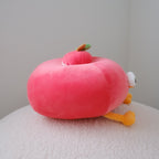 A pink plush apple-shaped toy with small yellow legs, a white eye, and a green-and-orange accent on top, resting on a textured white surface against a plain light gray wall.