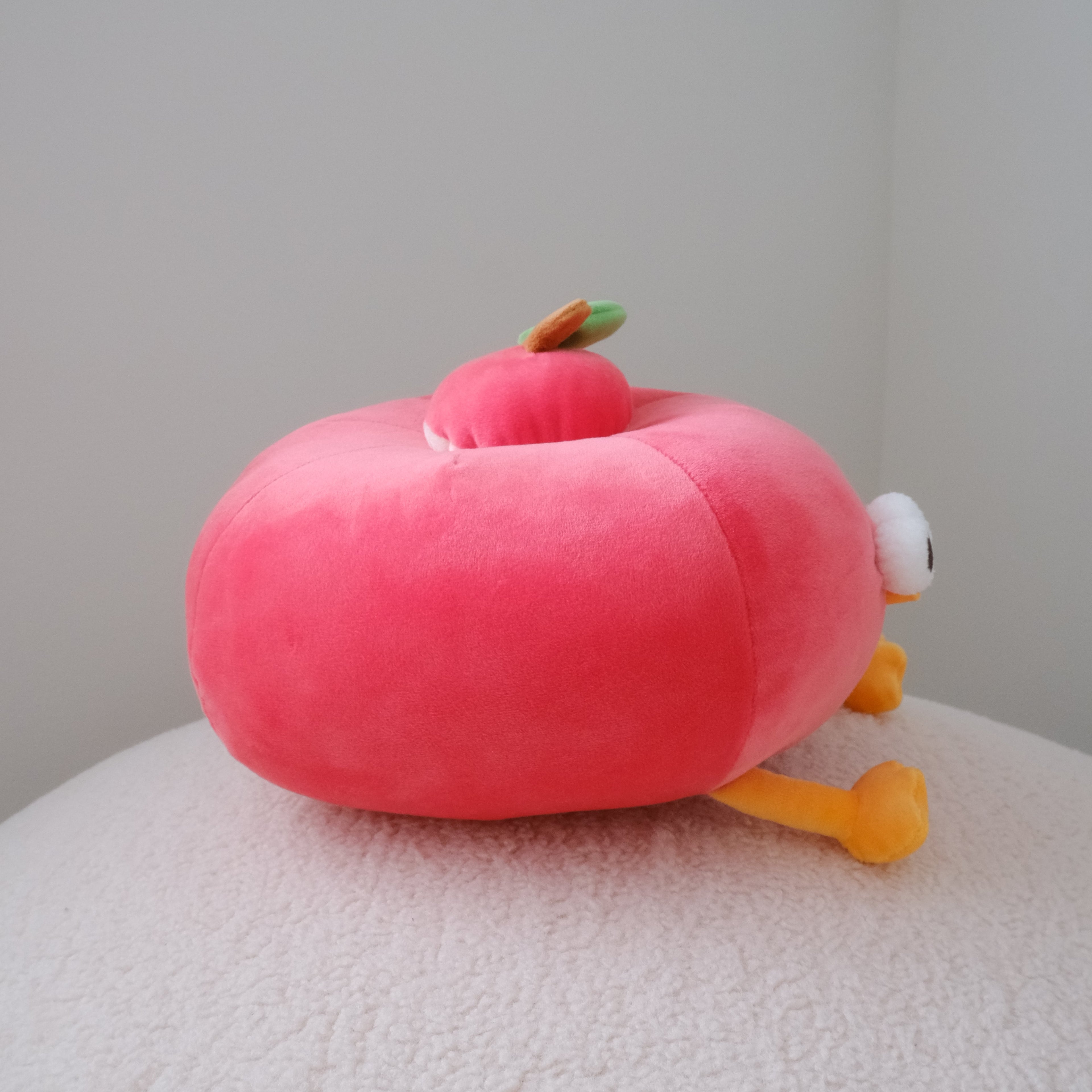 A pink plush apple-shaped toy with small yellow legs, a white eye, and a green-and-orange accent on top, resting on a textured white surface against a plain light gray wall.