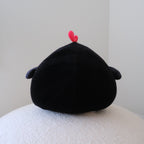 A black toy plush shaped chicken, viewed from the back, featuring a small red chicken crown on its head resting on a textured white surface against a plain light gray wall.