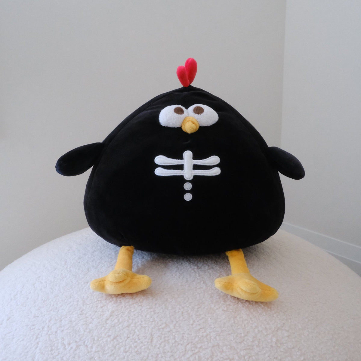 A black plush chicken toy with a red chicken crown, white embroidered eyes and white bones, and yellow beak and feet, sitting on a textured white surface against a plain light gray wall.