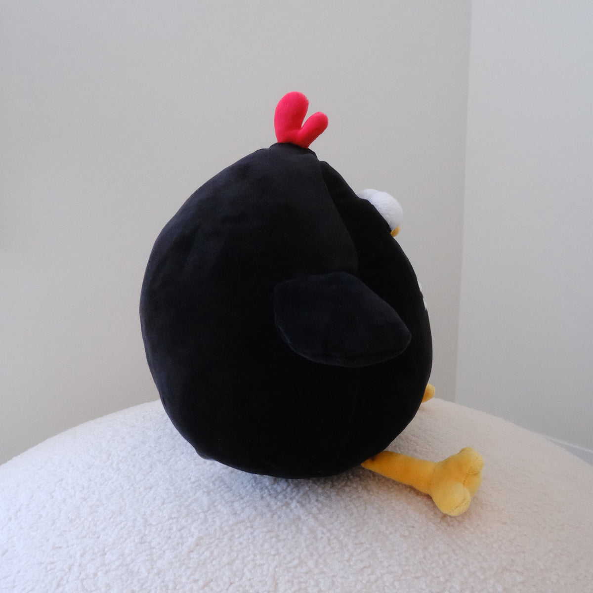 A black plush chicken toy with a red chicken crown, white eye, and yellow beak, sitting on a textured white surface against a plain light gray wall.
