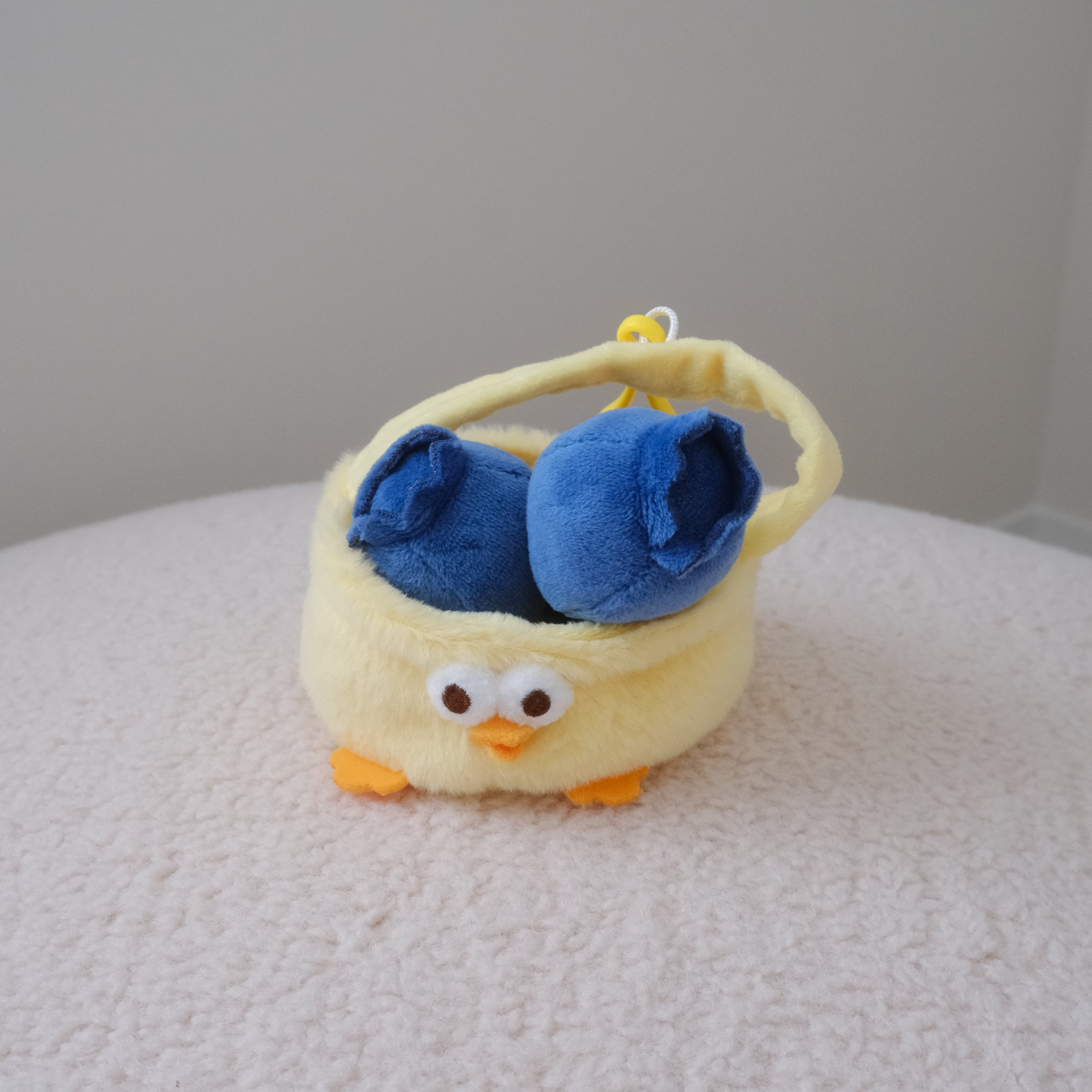 A small plush chicken basket in pale yellow with blueberry plush toys inside, white eyes with black pupils, and orange feet, placed on a textured white surface against a plain light gray wall.