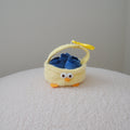 A small yellow plush basket with a chicken face and beak, holding three plush blueberries inside, set on a textured white surface against a plain light gray wall.