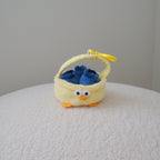 A small yellow plush basket with a chicken face and beak, holding three plush blueberries inside, set on a textured white surface against a plain light gray wall.