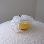 A back of a yellow chicken plush wearing a white tulle wedding dress and attached to a white ruffled wristlet strap, displayed on a textured cream fabric surface.