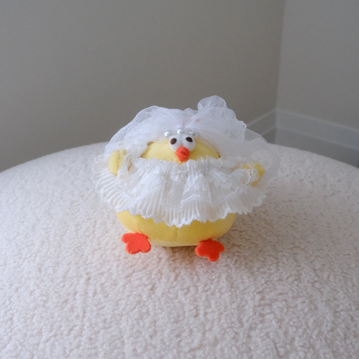 A small yellow plush chick toy wearing a white lace bridal veil and ruffled white dress, with orange feet and a small orange beak, standing on a textured white surface against a plain light gray wall.