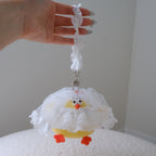 A hand holds a small yellow chick-shaped charm or bag with a white ruffled dress and orange feet against a plain indoor background.