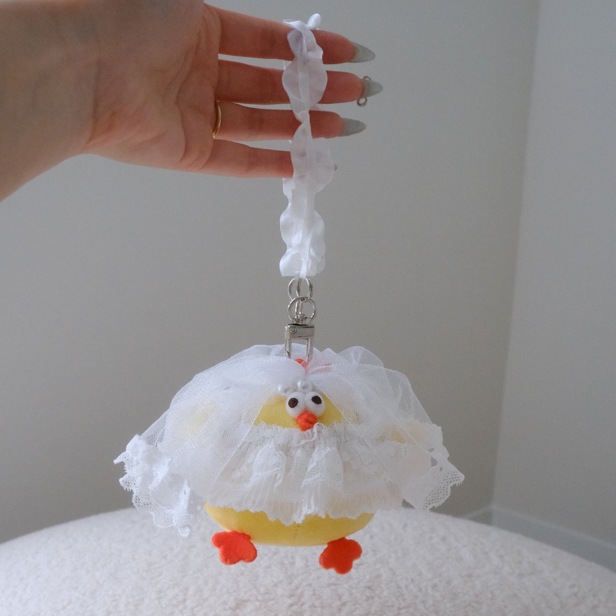 A hand holds a small yellow chick-shaped charm or bag with a white ruffled dress and orange feet against a plain indoor background.