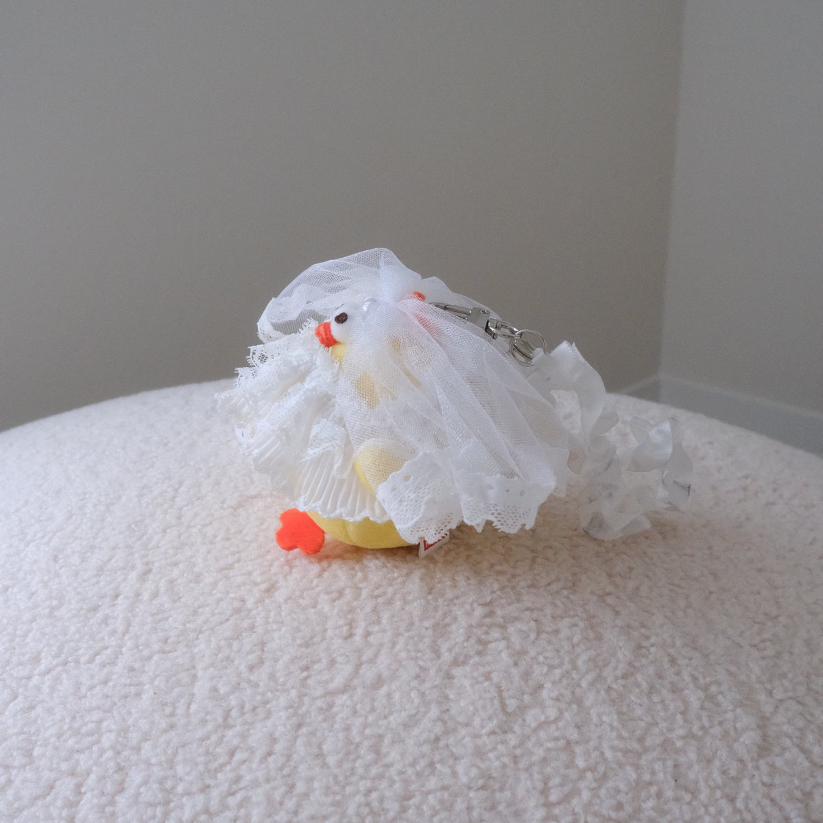 The side view of a yellow chicken plush wearing a white tulle wedding dress and attached to a white ruffled wristlet strap, displayed on a textured cream fabric surface.