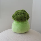 A plush green broccoli-shaped plush toy with a textured, shaggy green top and a smooth light green base, sitting on a round white textured surface against a plain light gray wall.