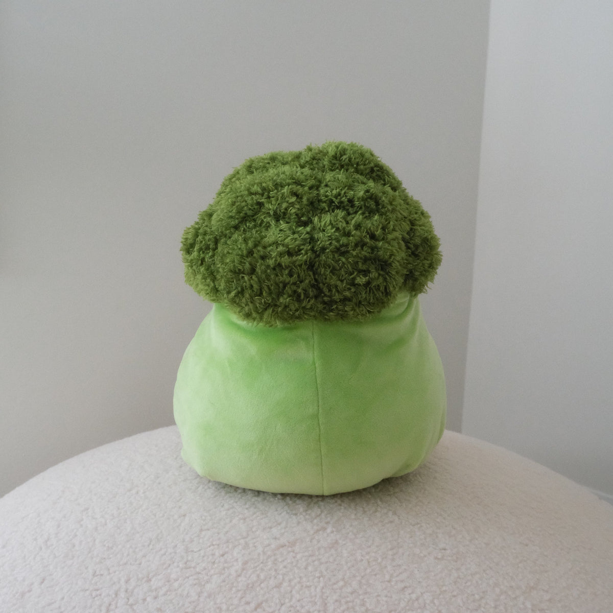 A plush green broccoli-shaped plush toy with a textured, shaggy green top and a smooth light green base, sitting on a round white textured surface against a plain light gray wall.