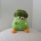 A green broccoli-shaped plush toy with orange feet and a white, black, and orange face, sitting on a textured white surface against a plain light gray background.