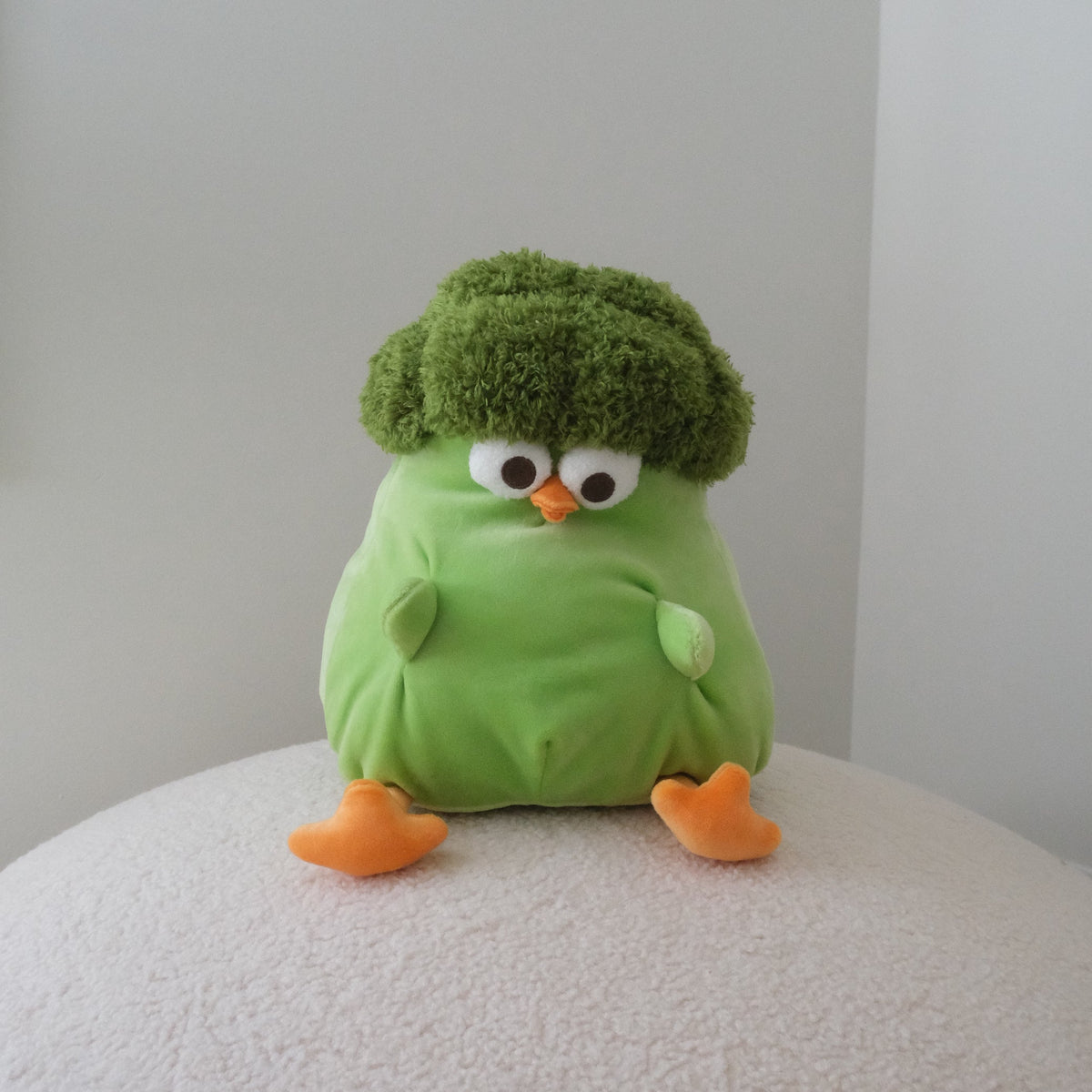 A green broccoli-shaped plush toy with orange feet and a white, black, and orange face, sitting on a textured white surface against a plain light gray background.