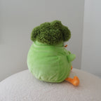 A plush toy shaped like a broccoli vegetable, seen from behind, sitting on a textured white surface against a plain light gray wall.
