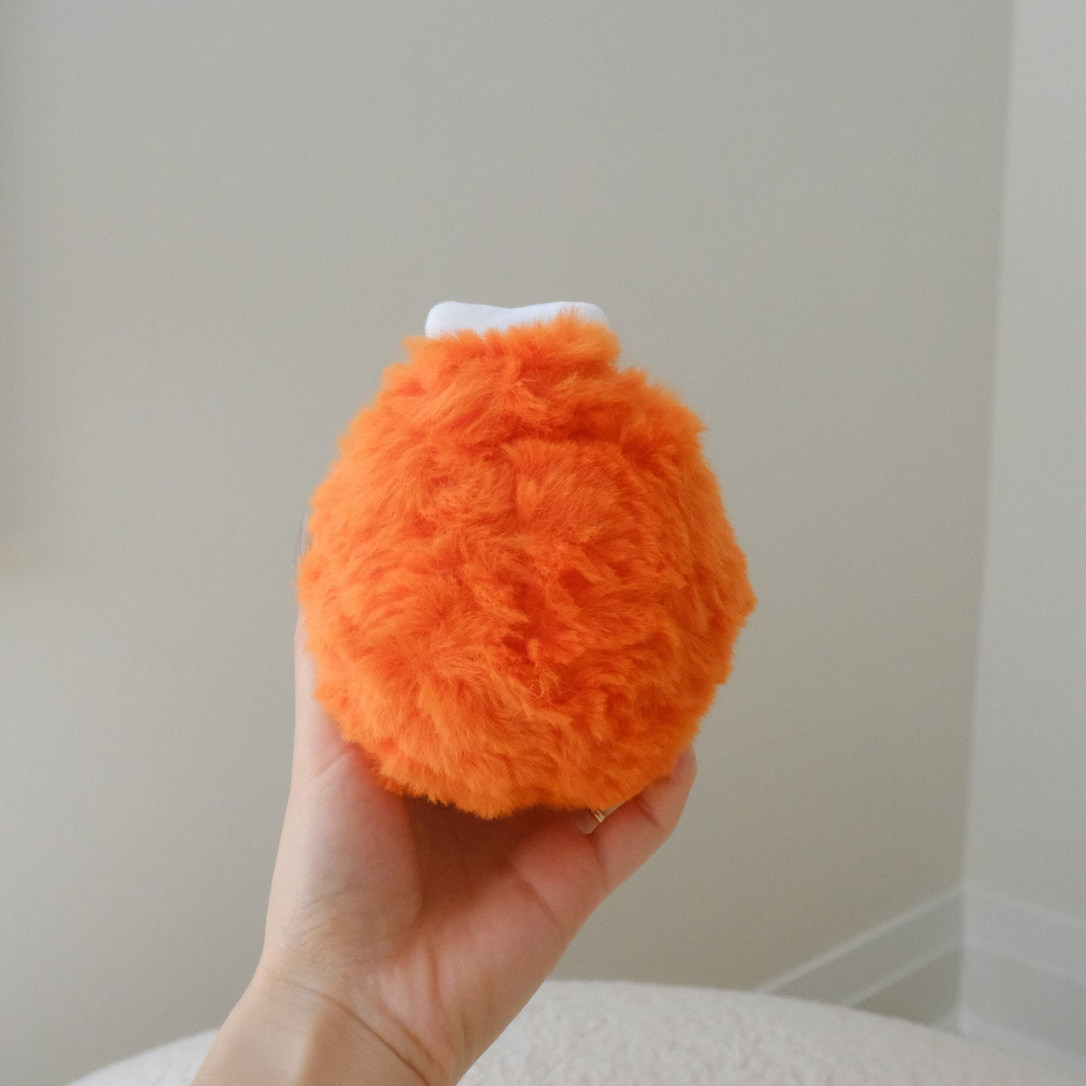 A hand holding a small, round, fluffy orange drumstick plush toy with a tiny white bone at the top, set against a soft-focus indoor background.