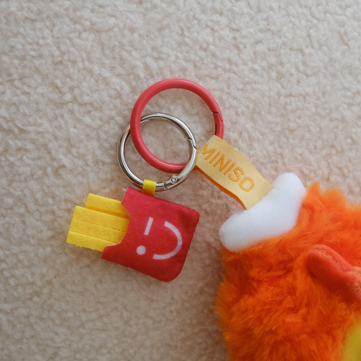 A close-up of a Miniso keychain charm shaped like a red french fry bag with yellow fries and a red key ring, shown next to an orange plush toy on a beige textured background.